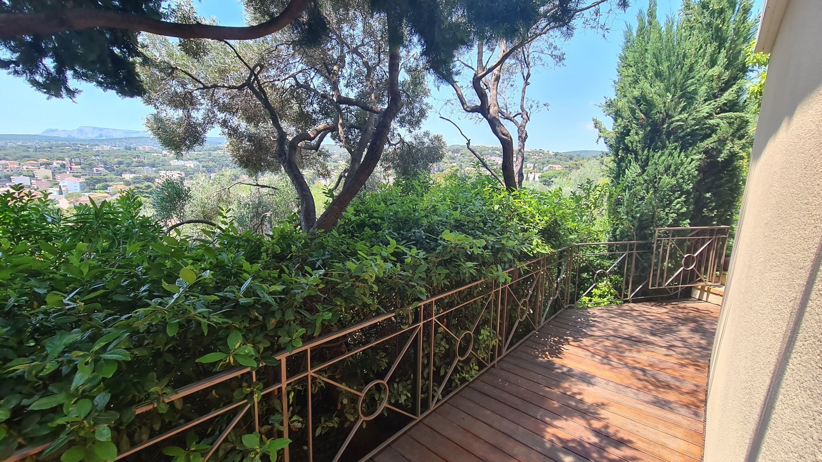 Une terrasse en bois avec une rambarde en métal, entourée d’une végétation luxuriante et offrant une vue sur le paysage urbain.