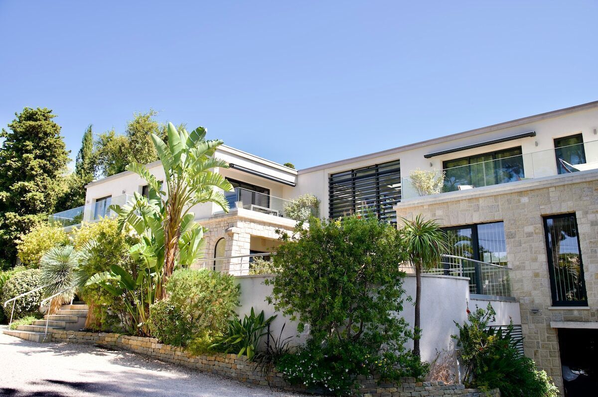 Una casa moderna de dos pisos con fachada de piedra, ventanas amplias y un jardín bien cuidado con diversas plantas y una palmera.