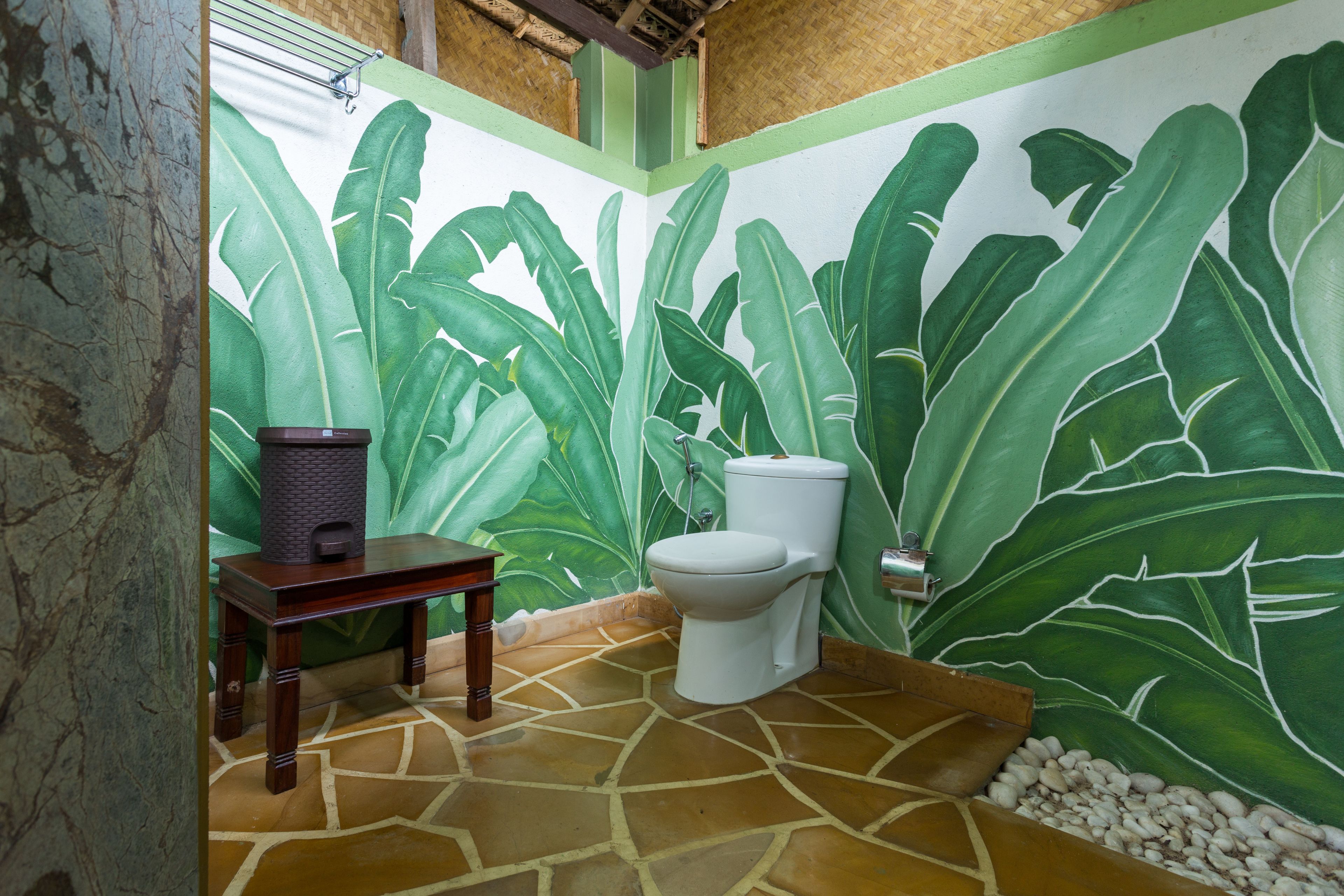A bathroom with a toilet, a small wooden table, and a large green leaf mural on the wall.