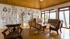 Une pièce avec un plafond haut, un ventilateur de plafond, une grande fresque murale, un canapé, deux fauteuils et une table en bois.