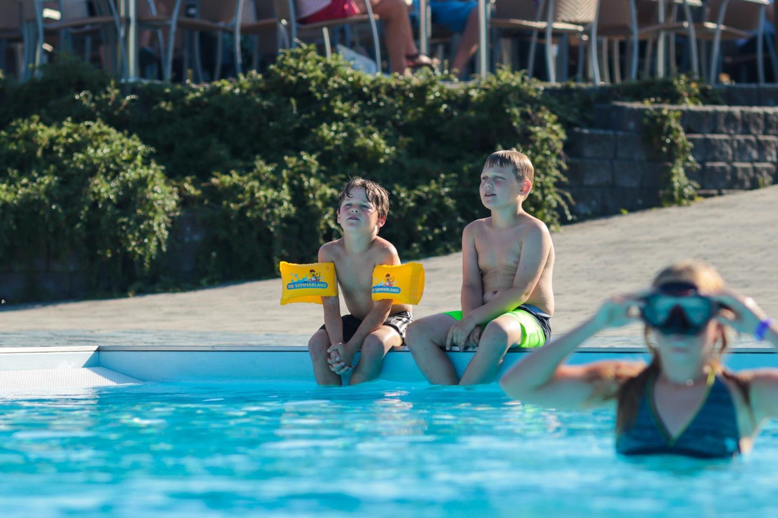 Deux personnes sont assises au bord d’une piscine, avec un sauveteur en arrière-plan.
