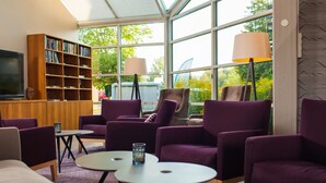 A modern living room with purple armchairs, a wooden bookshelf, a flat-screen TV, and large windows offering a view of greenery.