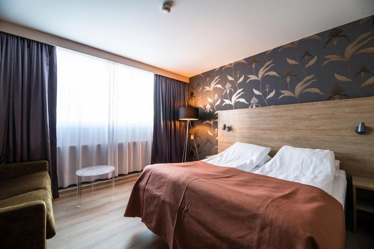 Quarto de hotel com cama, poltrona, mesa pequena, abajur e janela com cortinas.