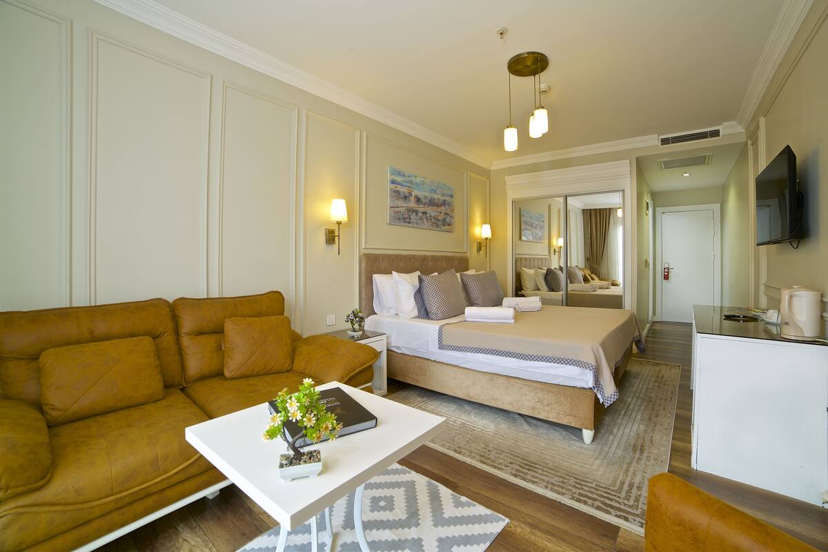 A hotel room with a brown leather sofa, a white coffee table with a book and flowers, a bed with a white and blue checkered bedspread, a bedside table with a lamp, and a television on the wall.