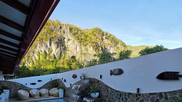 Una piscina con pared de piedra y una cascada, rodeada de mobiliario de mimbre y plantas en macetas.