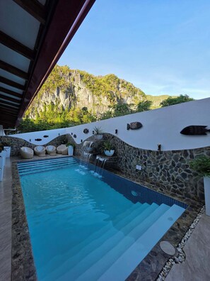 Una piscina con pared de piedra y una cascada, rodeada de mobiliario de mimbre y plantas en macetas.
