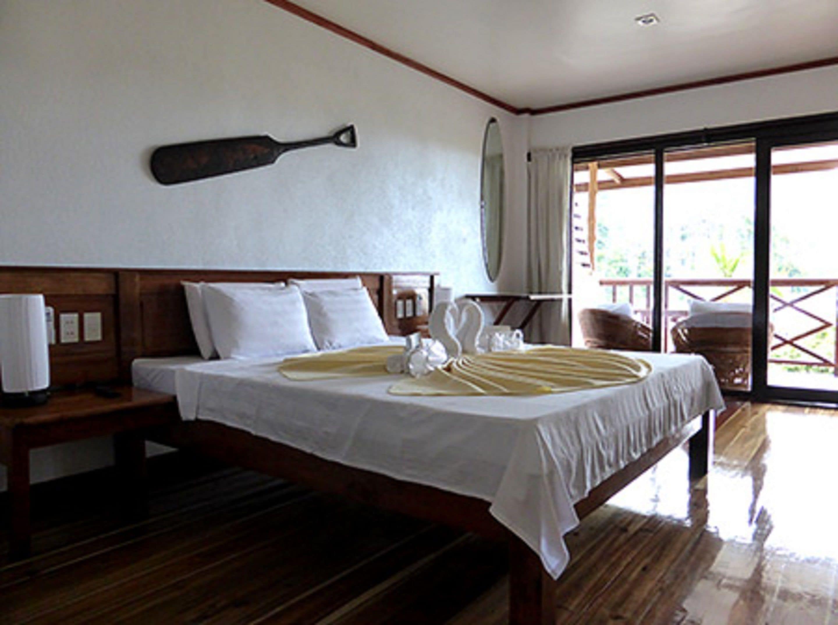 Habitación de hotel con una cama de madera, ropa de cama blanca, una mesita de noche con lámpara y vistas al exterior a través de una puerta corrediza de vidrio.
