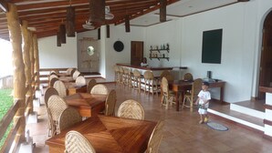 Un amplio comedor con mesas de madera y sillas de mimbre, una zona de bar con estantes llenos de botellas, y vistas al exterior.