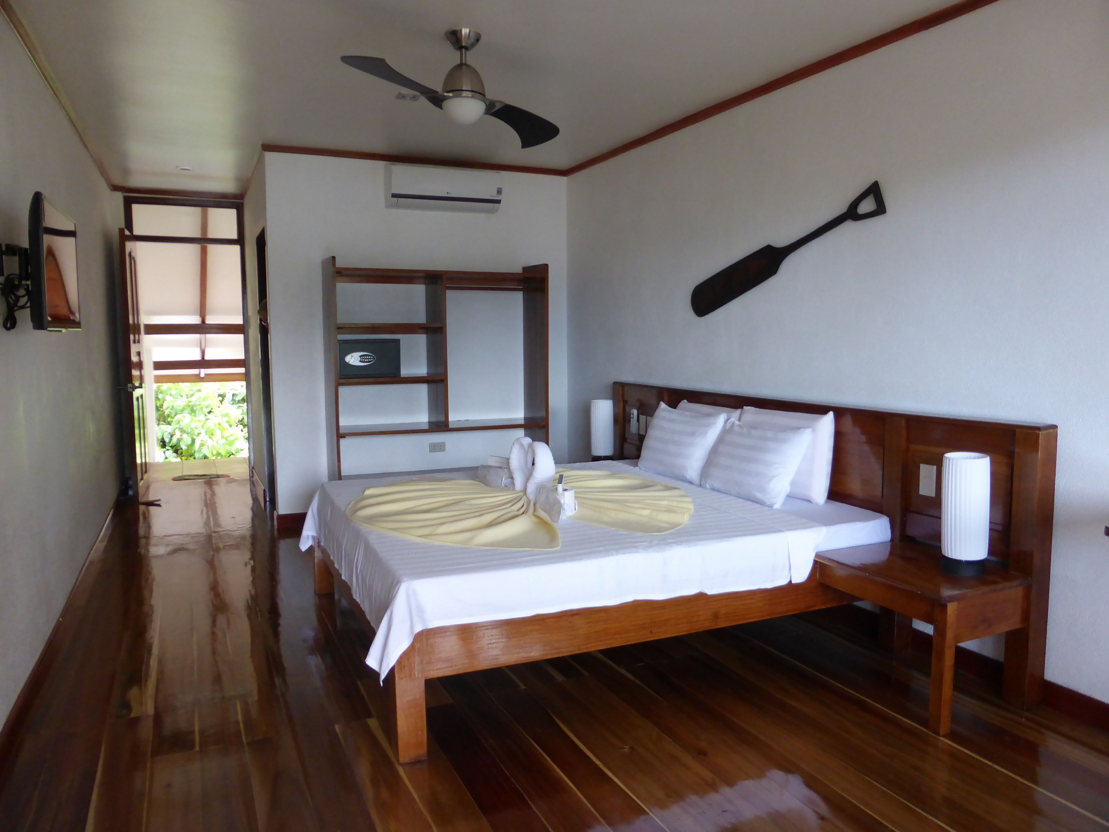 A hotel room with a wooden bed, a ceiling fan, a wooden headboard, a bedside table, and a view of greenery outside.