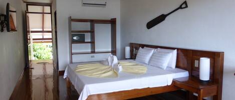 A hotel room with a wooden bed, a ceiling fan, a wooden headboard, a bedside table, and a view of greenery outside.