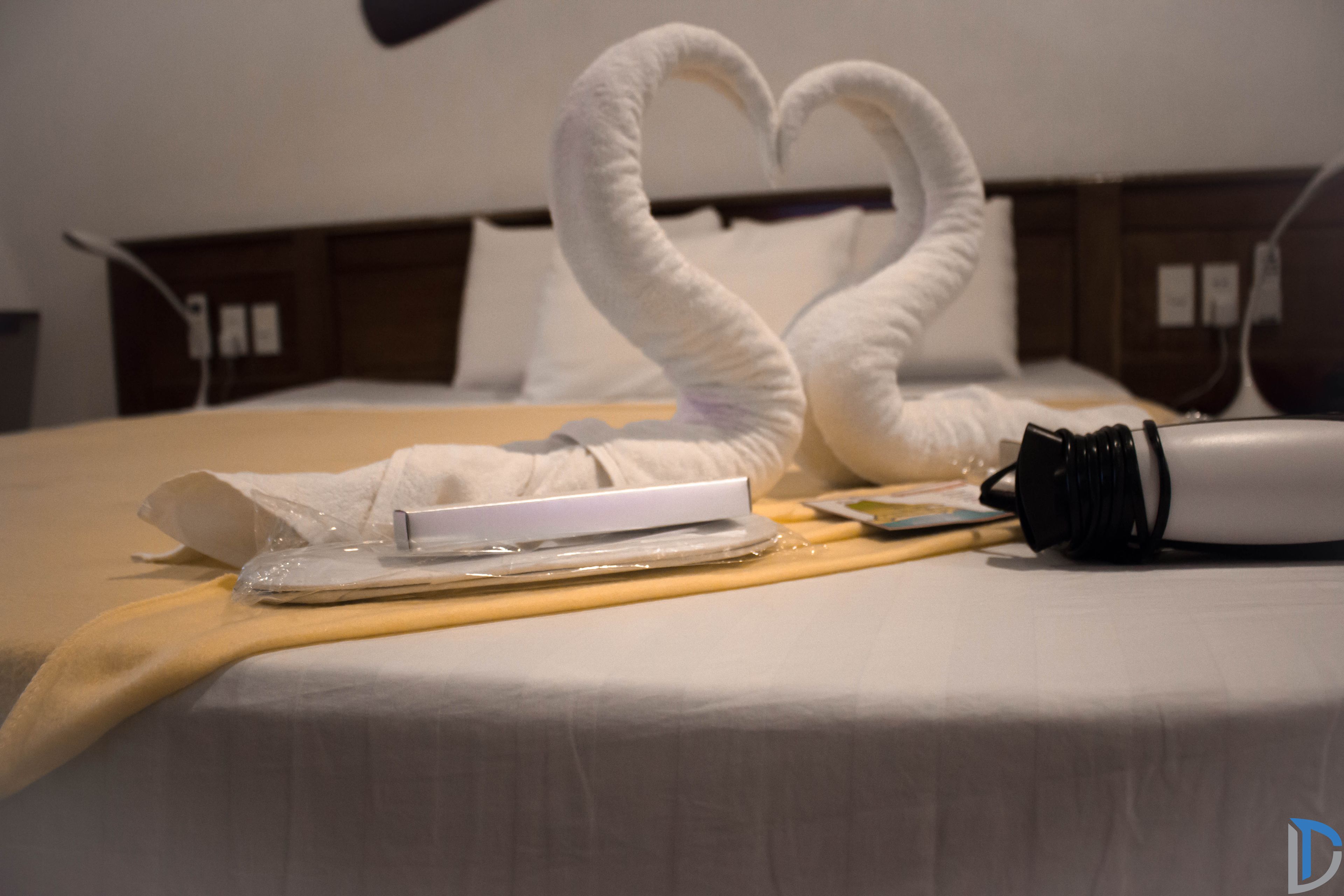 A neatly made bed with a heart-shaped towel arrangement, a remote control, a bottle, and a tissue box.