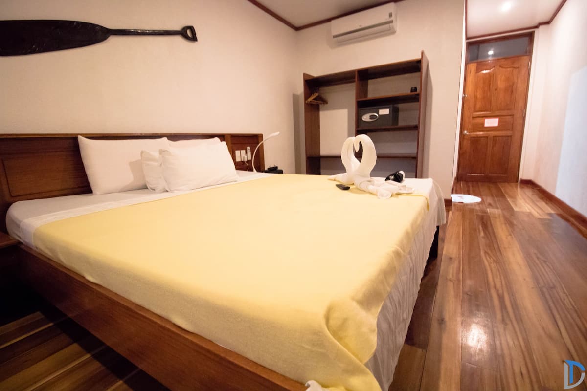 Un letto rifatto con una coperta gialla, una testiera in legno e un cuscino bianco. Si nota un mobiletto a mensole con un asciugamano e un porta asciugamani a forma di cigno bianco sul letto.