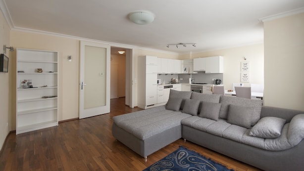 A modern living room with a grey sectional sofa, a wooden floor, and a kitchen in the background. - Heart of Vienna Residence (Vienna)