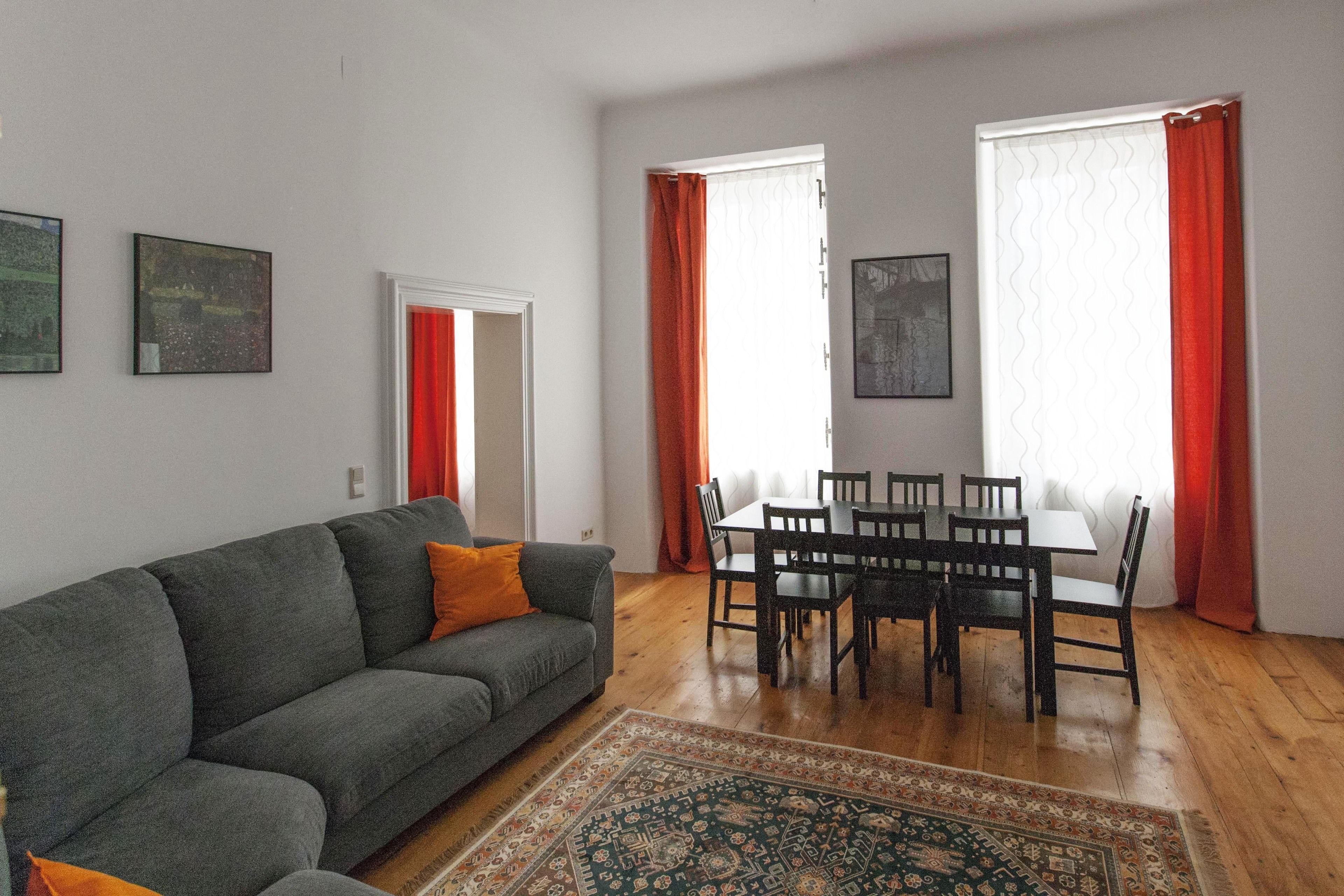A living room with a grey sofa, a patterned rug, a dining table with chairs, and two windows with red curtains.