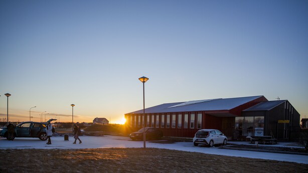 En bygning med en stor glasfacade og rødt ydre, beliggende i et snækkedækket område med en solnedgang i baggrunden.