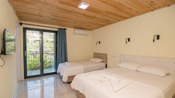 A hotel room with two beds, a balcony with a view, wooden ceiling, and a flat-screen TV. - Beyoglu Pansiyon Apart (Kas)