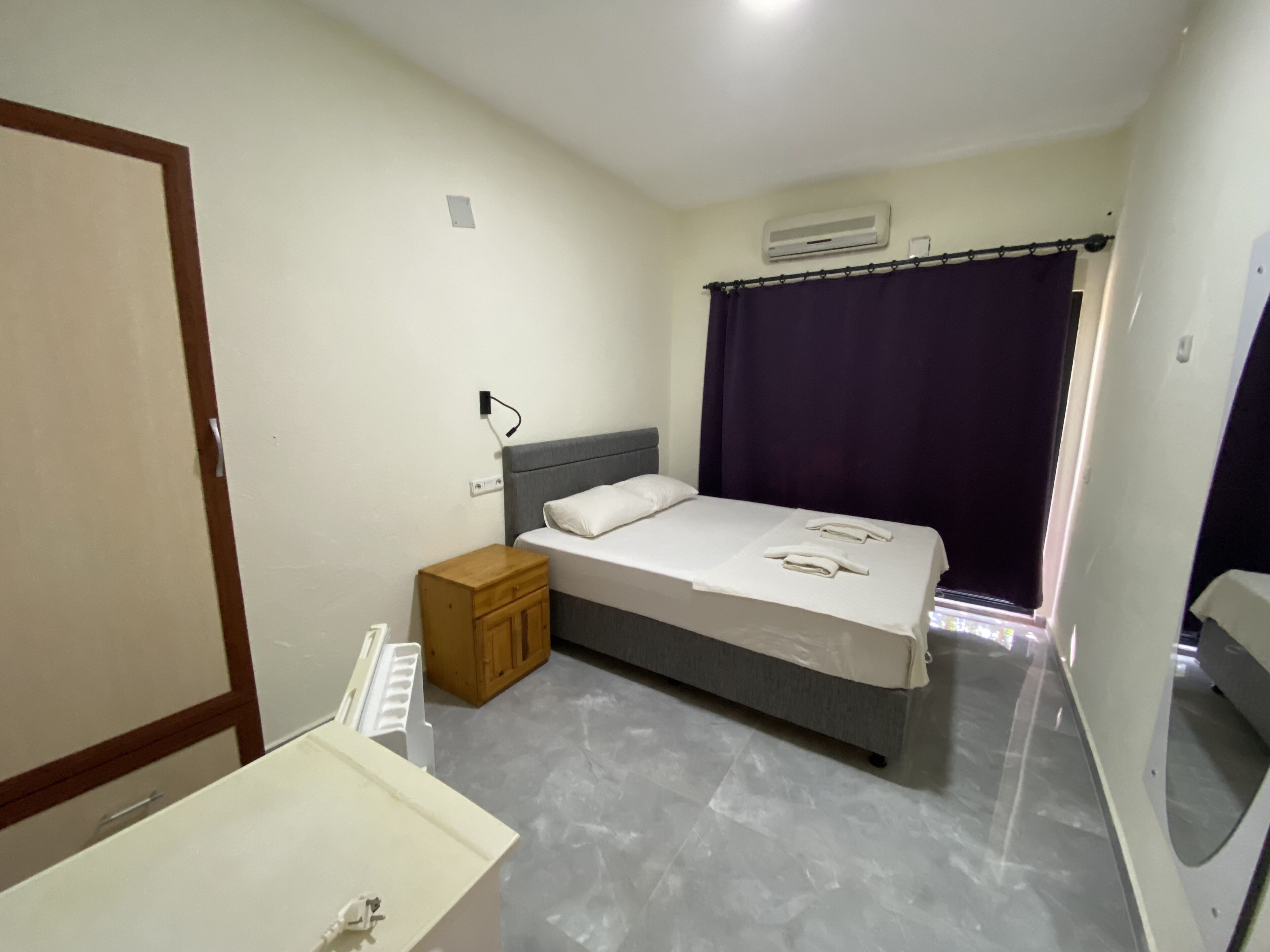 A hotel room with a bed, a nightstand, a wooden cabinet, a bathroom with a mirror, and an air conditioning unit.