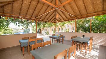 A covered outdoor dining area with wooden tables and chairs, a thatched roof, and a view of greenery and buildings in the background.