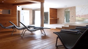 A modern spa room with wooden floors, two lounge chairs, a stone wall, and a glass door leading to a pool area.