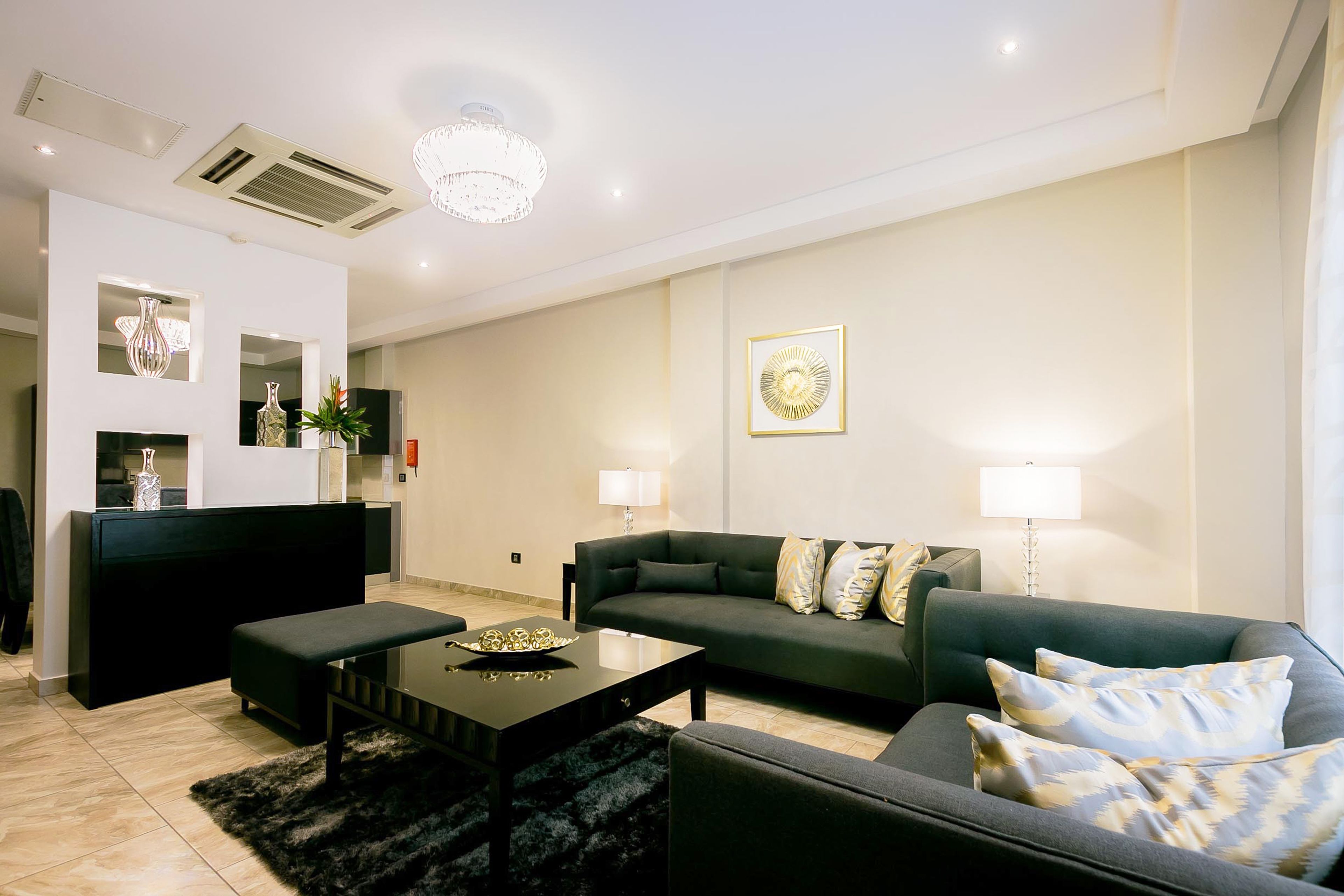 Una sala de estar moderna con un sofá, una mesa de centro y un reposapiés negro. Hay un televisor sobre un mueble, un espejo decorativo y una araña.