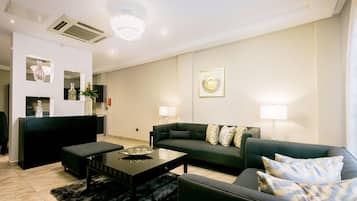 Una sala de estar moderna con un sofá, una mesa de centro y un reposapiés negro. Hay un televisor sobre un mueble, un espejo decorativo y una araña.