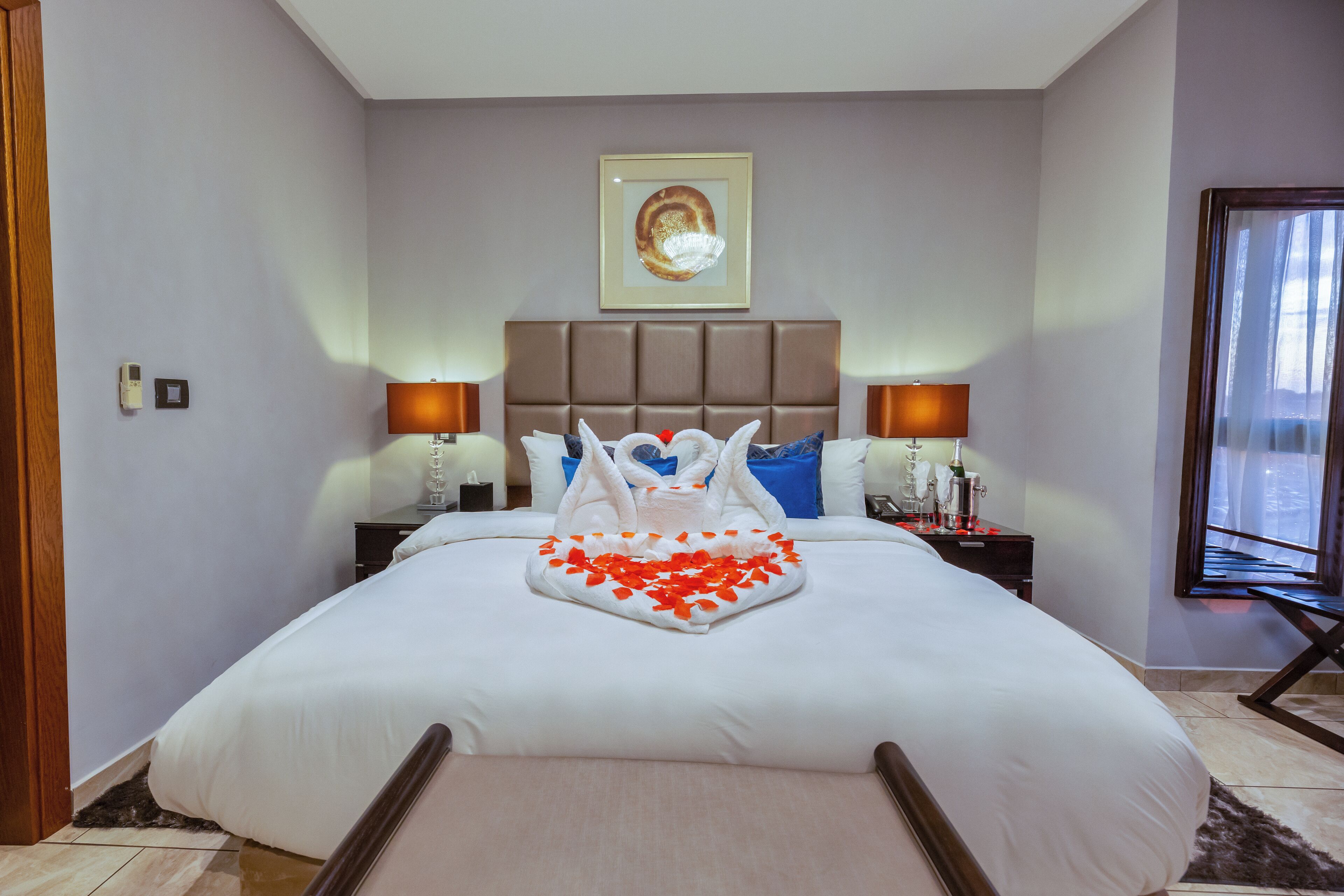 Een hotelkamer met een groot bed, een hoofdeinde, nachtkastjes met lampen, een lijst met een afbeelding aan de muur, en een raam met gordijnen.