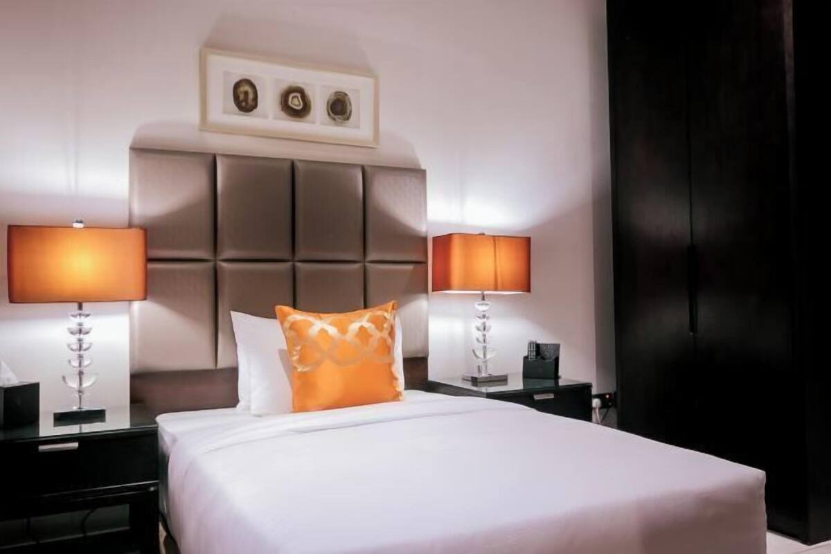 A hotel room with a bed, two bedside lamps, a nightstand, a framed picture above the bed, and a dark door.