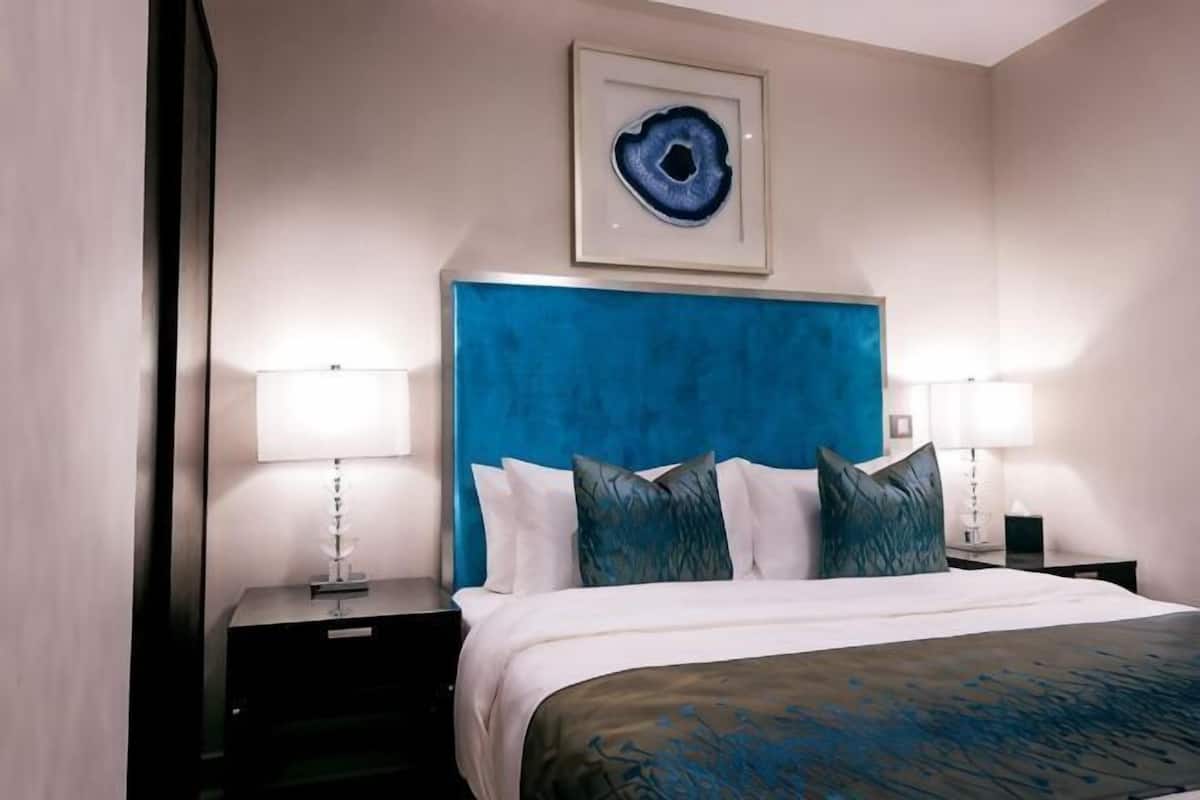 A hotel room with a large bed, two bedside tables, a blue headboard, and a framed artwork above the bed.