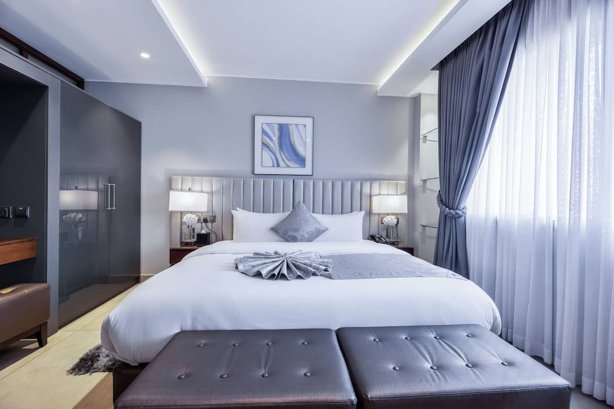 A modern hotel room with a large bed, two bedside tables with lamps, a grey upholstered bench, and a glass-enclosed walk-in closet.