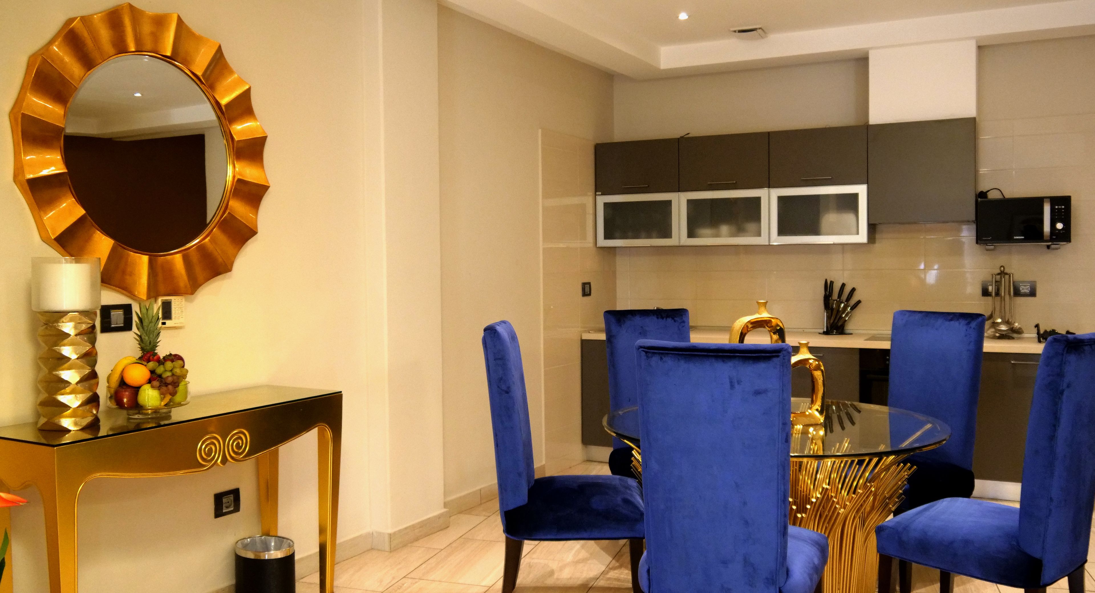 A modern dining area with a glass table, blue velvet chairs, and a gold-framed mirror.