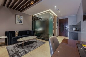 A modern living room with a dark blue sofa, a round coffee table, and a glass partition leading to a kitchen area.