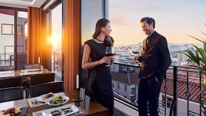 Een koppel dineert aan een tafel met uitzicht over de stad, een balkon en een ondergaande zon.
