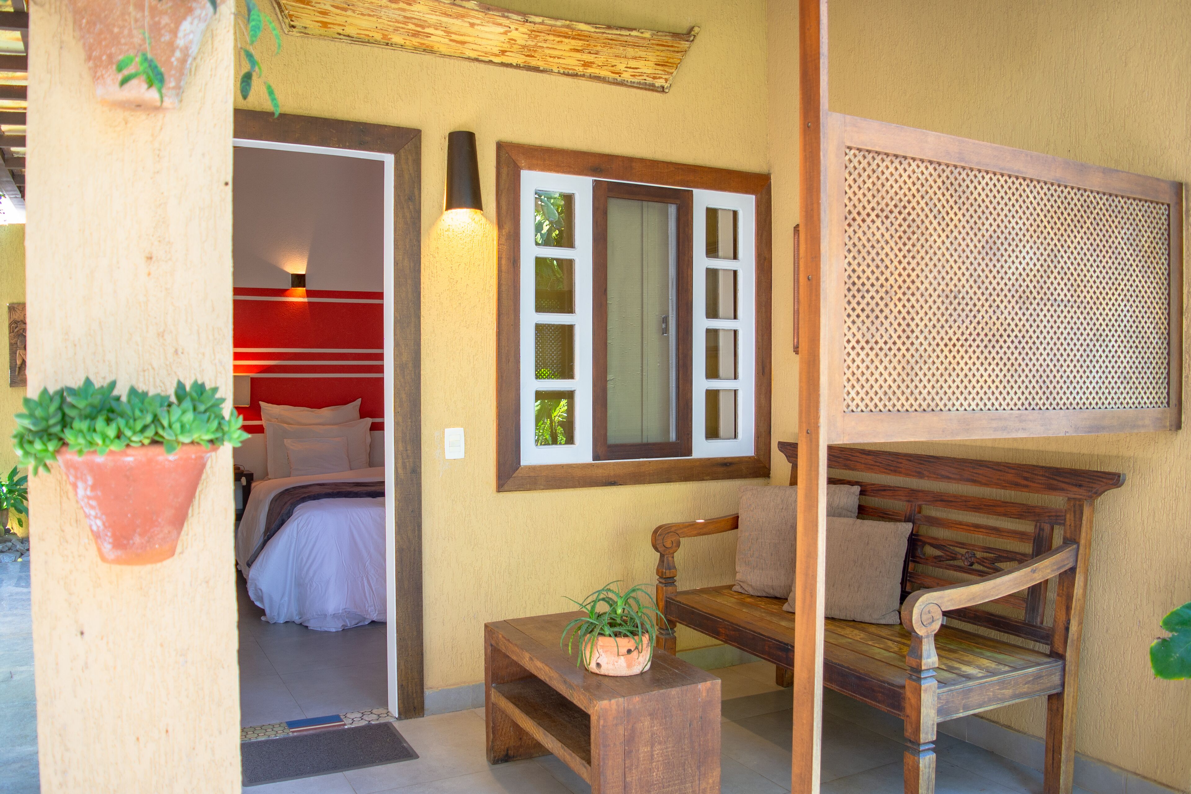 Una habitación con cama, un banco de madera, una mesita con una planta y un ventanal con marco de madera.
