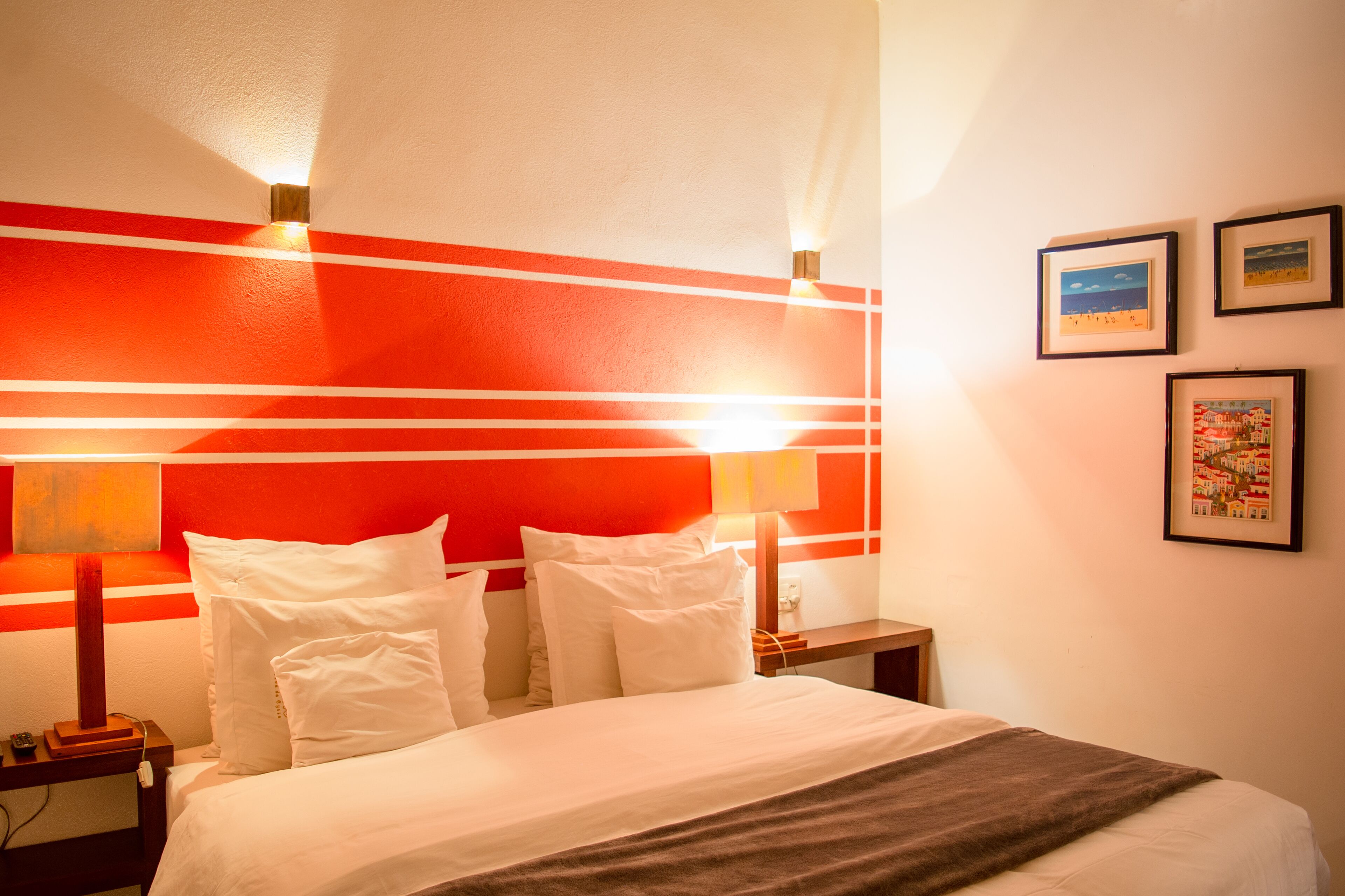 Quarto de hotel com cama, roupa de cama branca, parede de destaque vermelha e quadros nas paredes.