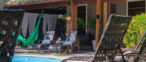 A poolside area with wicker lounge chairs, a hammock, and a tiled roof.