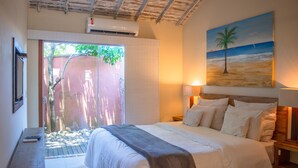 Ein Schlafzimmer mit Bett, Nachttischlampen, einem Bild mit Strandmotiv und Blick auf einen Garten durch eine Schiebetür.