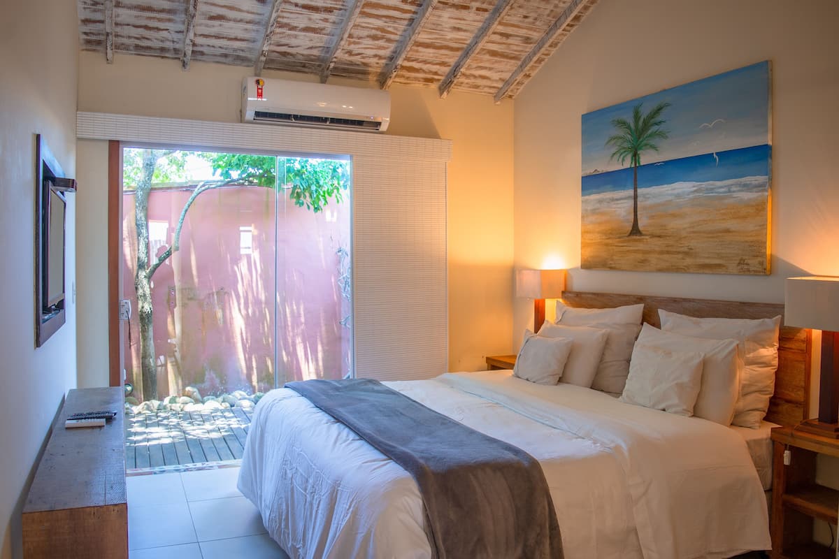 A bedroom with a bed, bedside lamps, a painting of a beach scene, and a view of a garden through a sliding door.