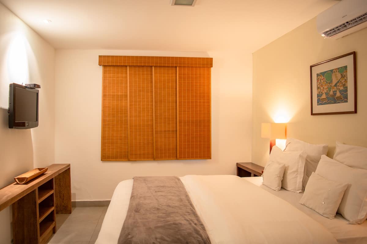 A bedroom with a bed, a television on a stand, a bedside table, a lamp, and a window with blinds.