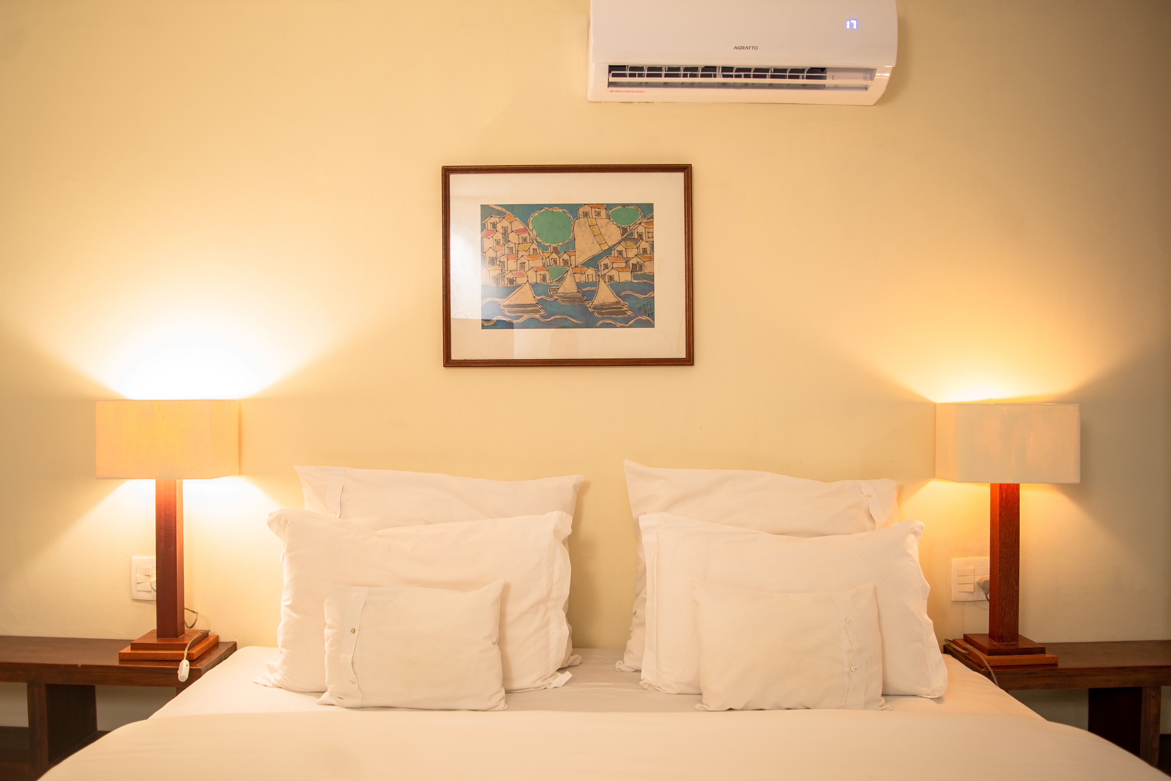 A hotel room with a bed, two bedside lamps, a framed picture on the wall, and an air conditioning unit above the bed.