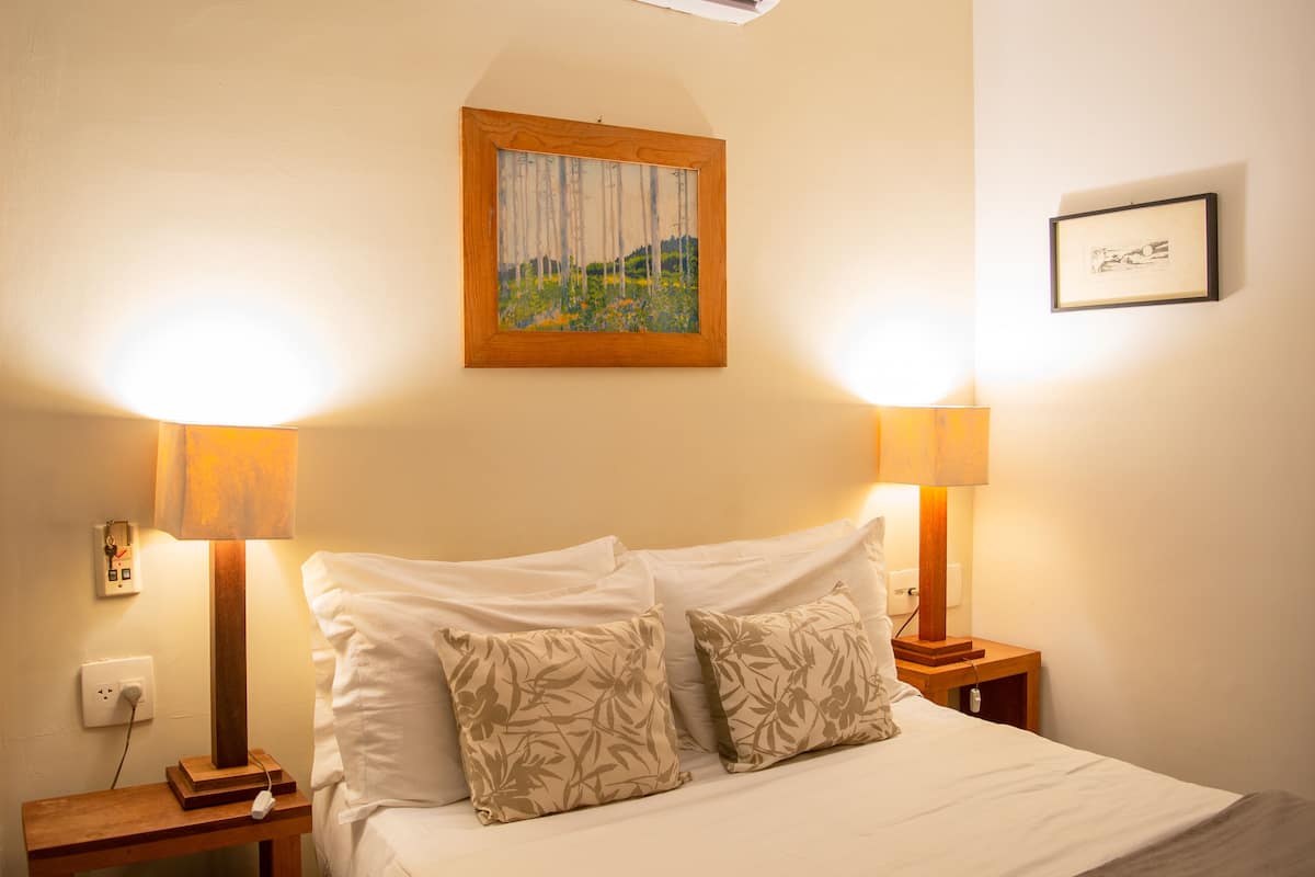A neatly made bed with floral-patterned pillows, two bedside lamps, a framed painting above the bed, and a small bedside table with a lamp.
