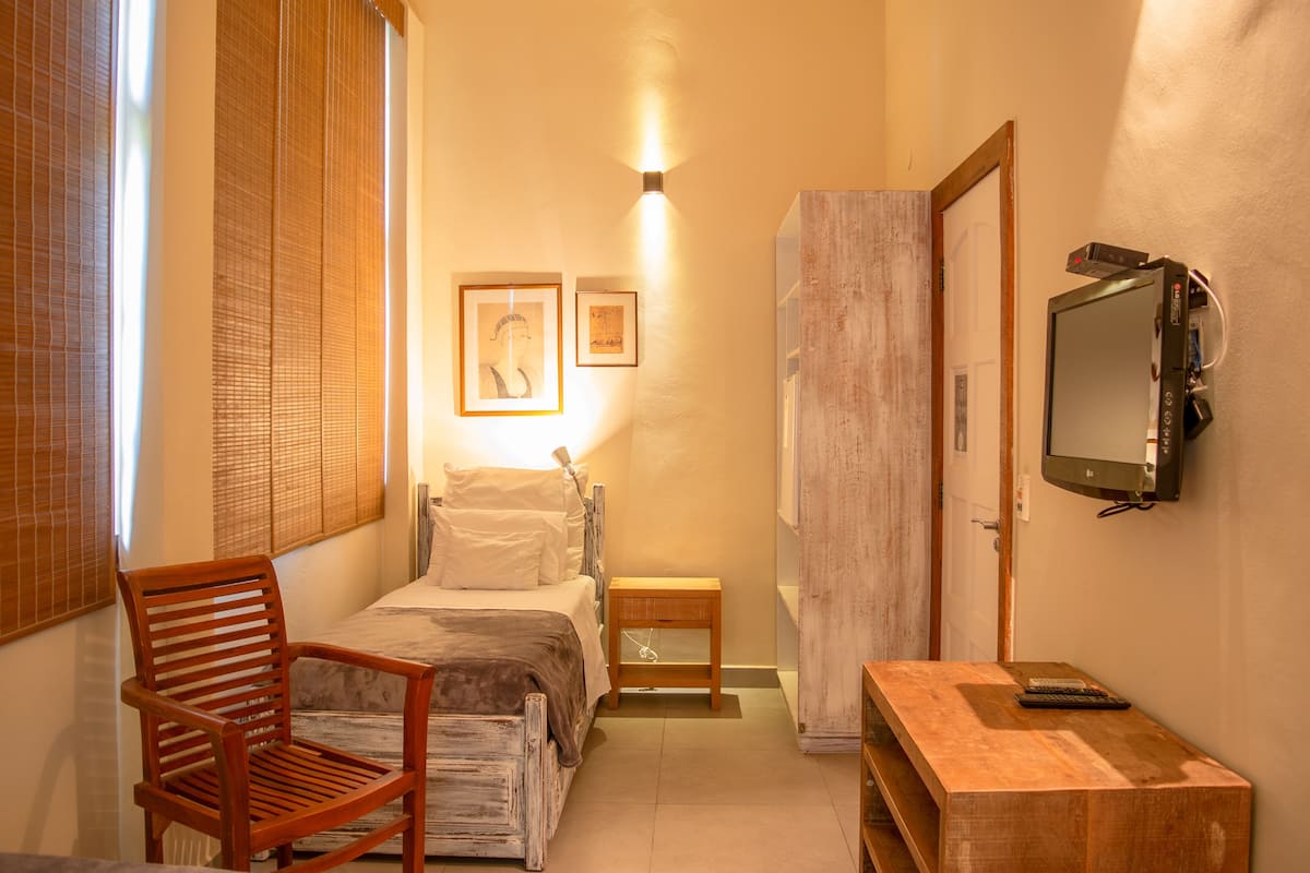 A narrow hotel room with a bed, a wooden chair, a small table with a remote control, and a television mounted on the wall.