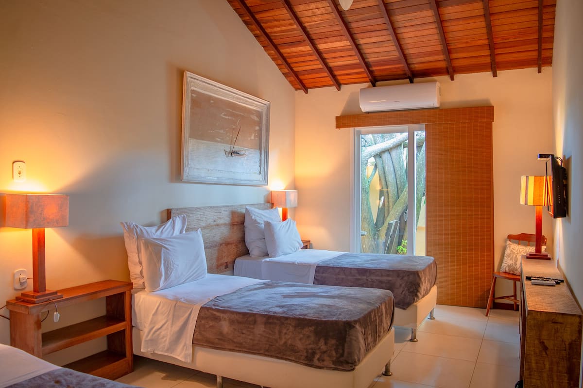 A hotel room with two beds, a wooden headboard, a desk with a lamp, and a large window with a view of greenery.