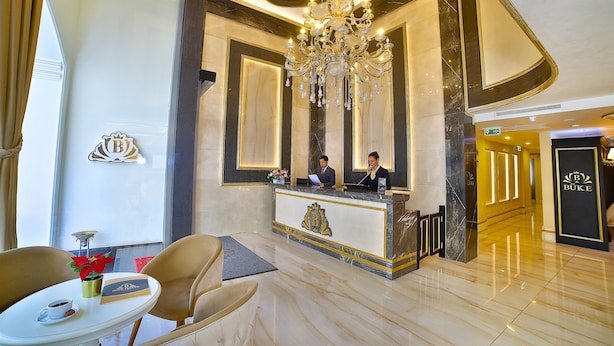 Zona de recepción de un hotel con un mostrador de mármol, una araña y dos recepcionistas.