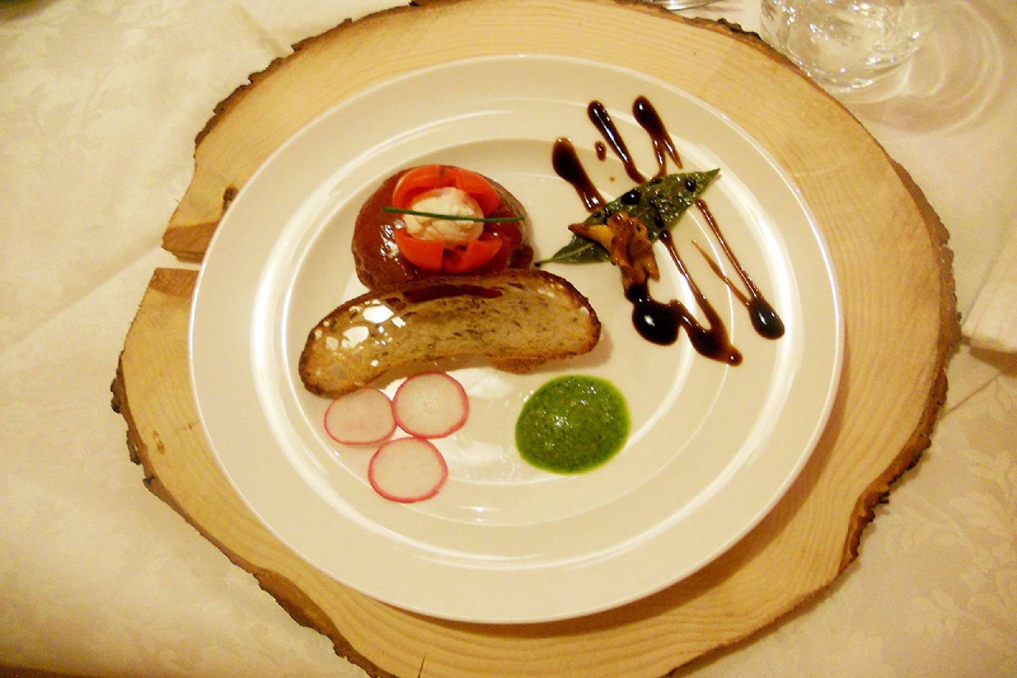 A plate with a sausage, tomato, radish, and green sauce on a wooden plate.