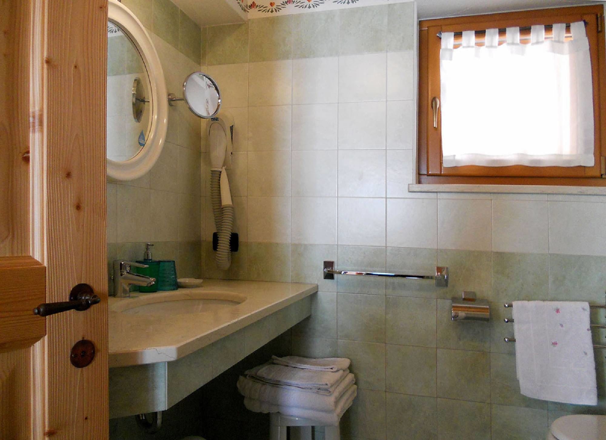 Une salle de bain avec une porte en bois, un miroir rond, un porte-serviettes, des toilettes et une fenêtre avec des rideaux.
