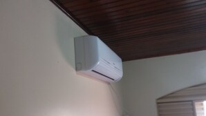 Een witte airconditioningunit bevestigd aan een muur in een kamer met een houten plafond en een raam met zonwering.