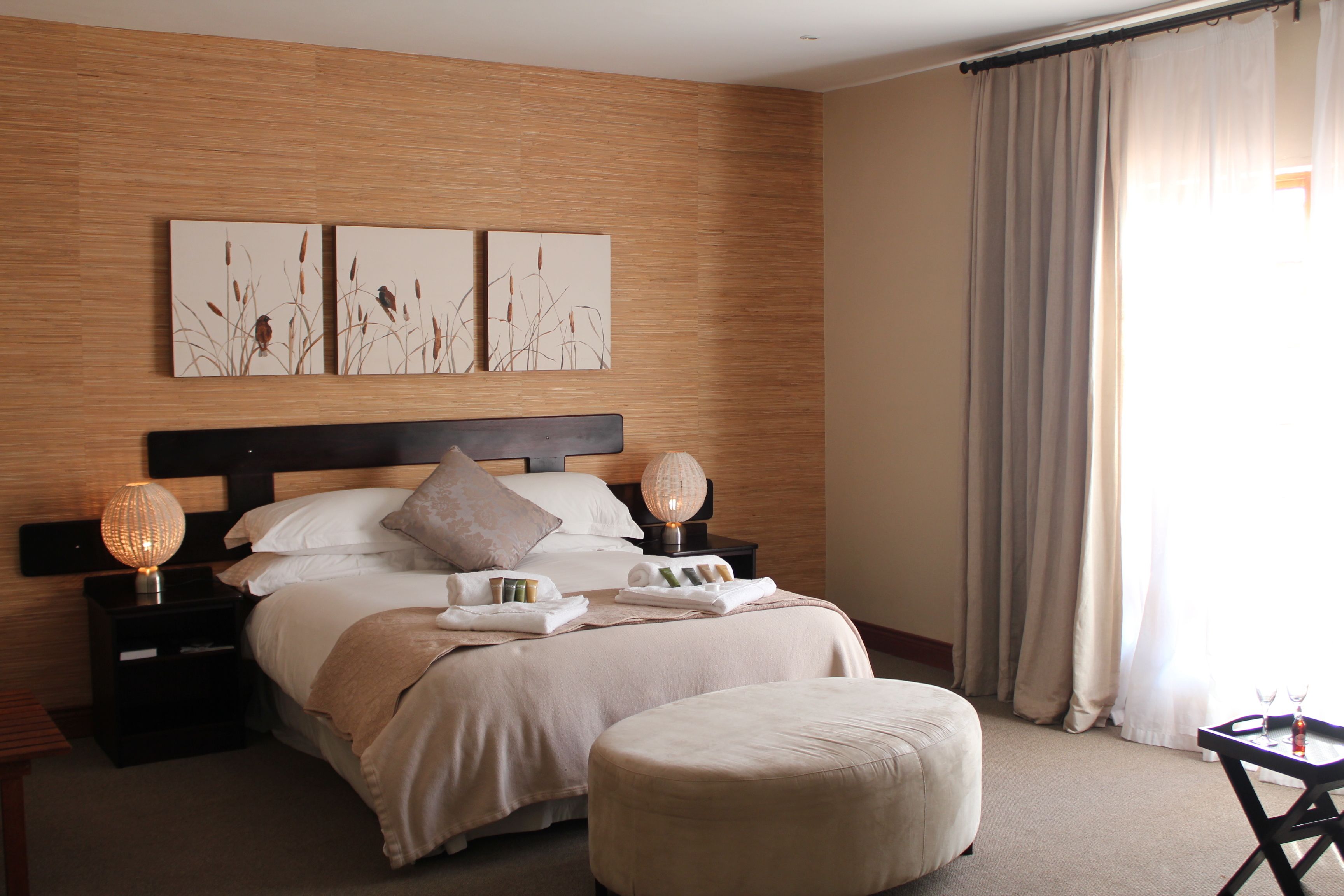 A hotel room with a wooden headboard, a bed with a beige bedspread, two bedside lamps, a nightstand with a lamp, a small table with a glass, and a window with curtains.
