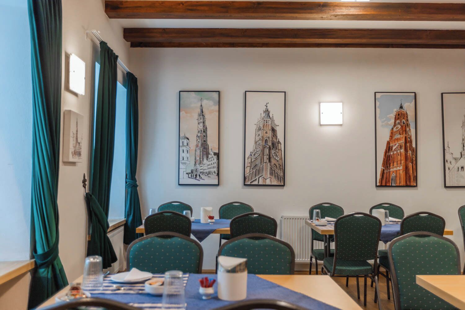 Een eethoek met tafels en stoelen, wanddecoratie bestaande uit afbeeldingen van gebouwen, en een raam met gordijnen.