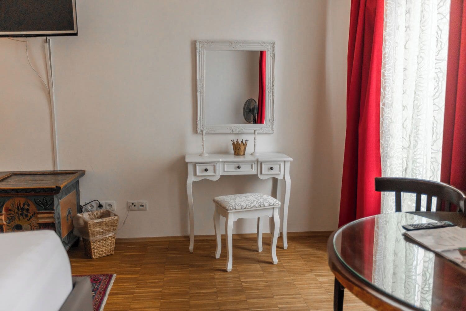 A room with a white vanity, a mirror, a small bench, a table with a chair, and a red curtain.