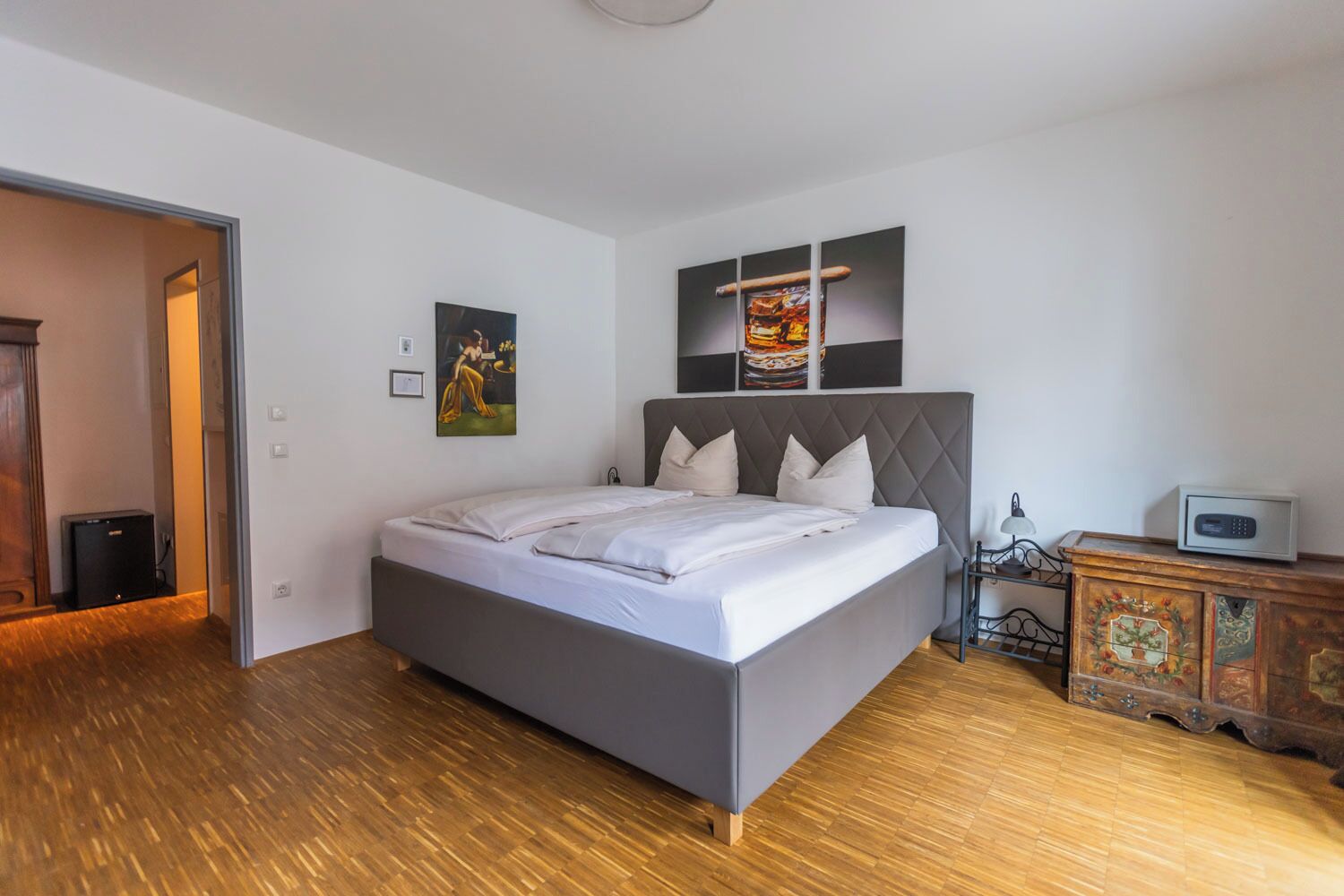 Un dormitorio moderno con una cama grande, suelo de madera, un aparador con paneles decorativos y un cuadro en la pared.